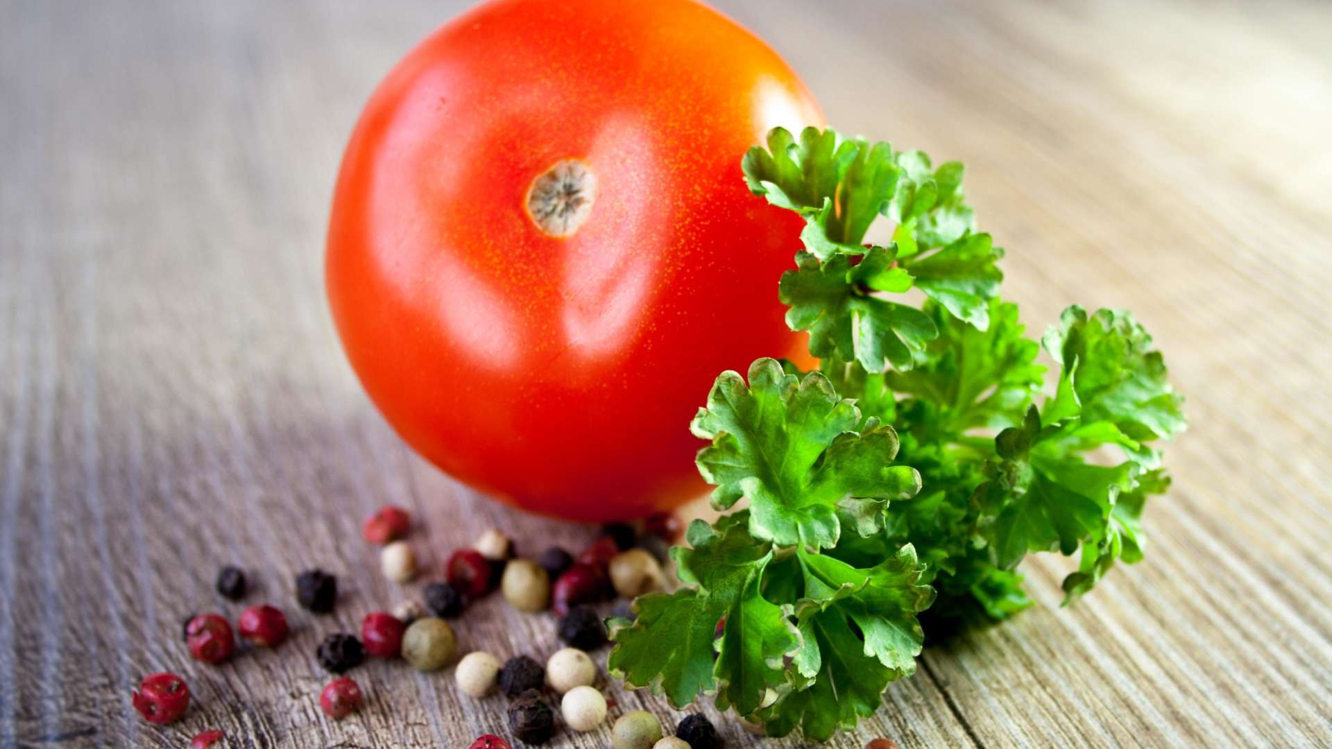 Punainen tomaatti, vihreätä salaattia ja erivärisiä pippureita pöydällä. Pippureita on mustia, punaisia ja valkoisia.