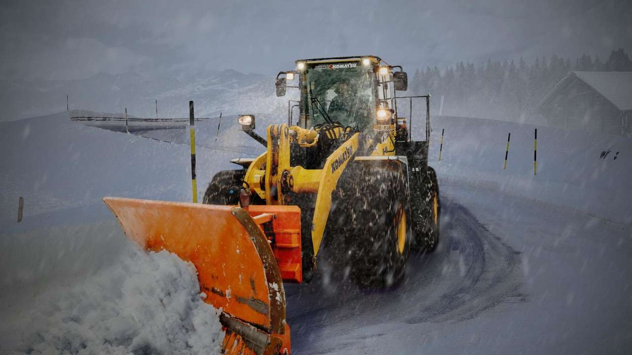 Keltainen traktori auraa tiellä lunta. On hämärää. Traktorissa on valot.