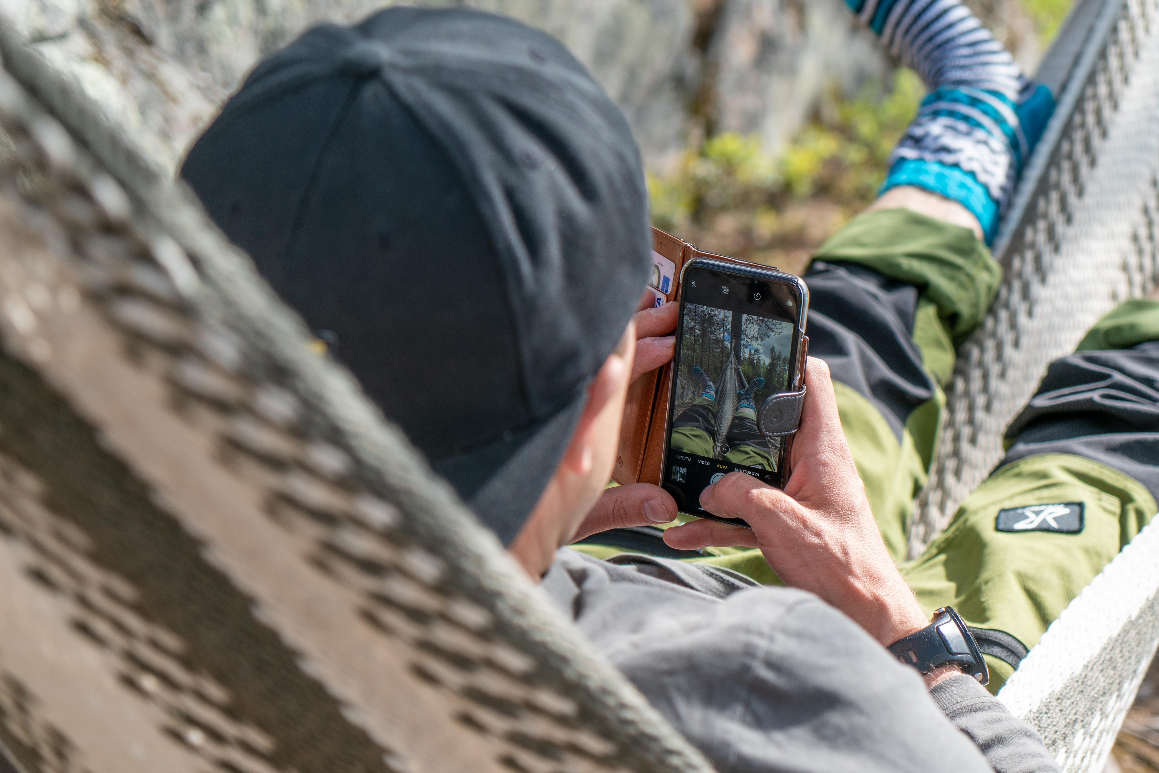 Mies Vuorijärvellä riippumatossa villasukat jalassa ottamassa kuvaa kännykällään.