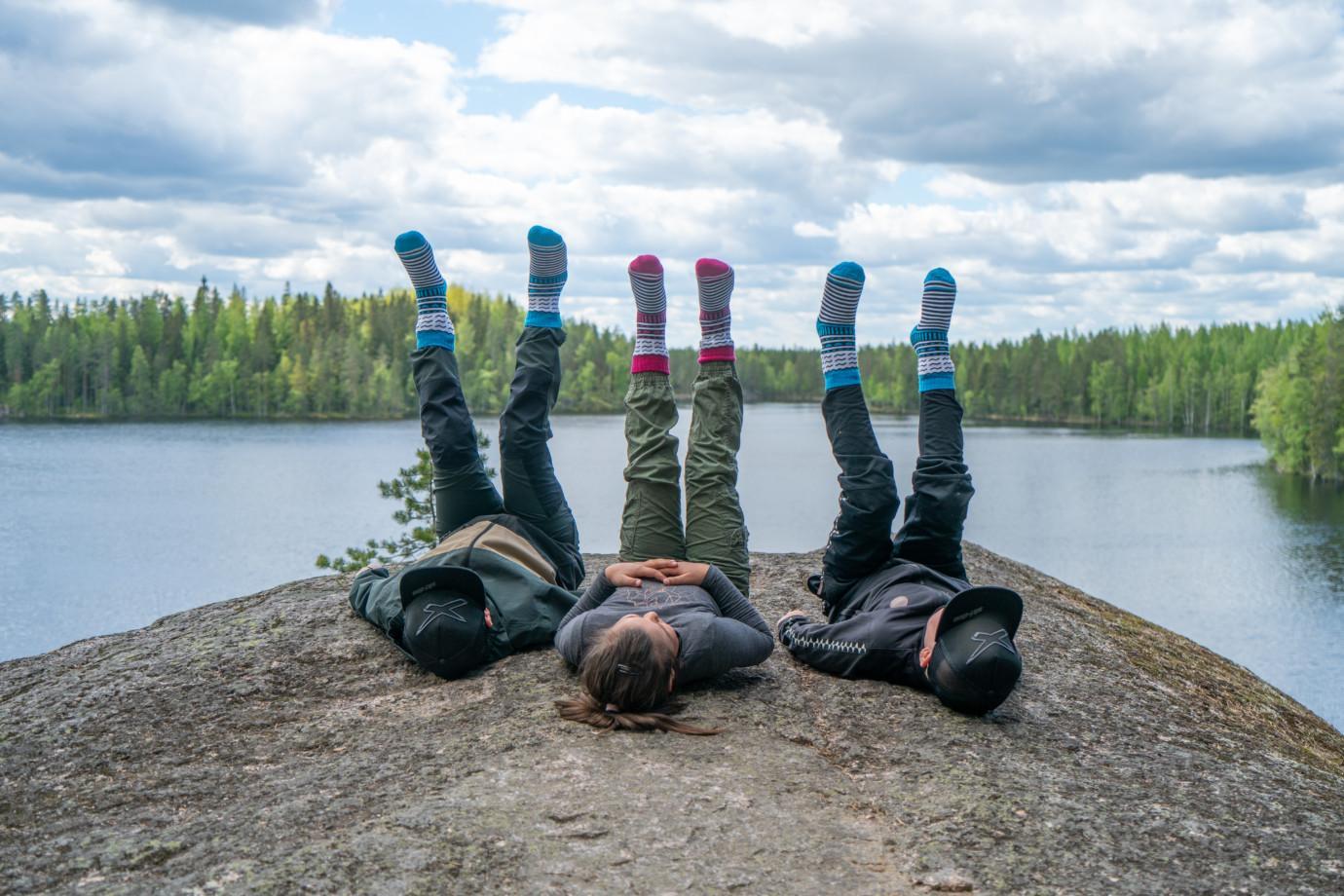 Kolme lasta makaa selällään kalliolla jalat kohti taivasta ja Orivesi-sukat jalassaan Iso-Vuorijärven maisemassa.
