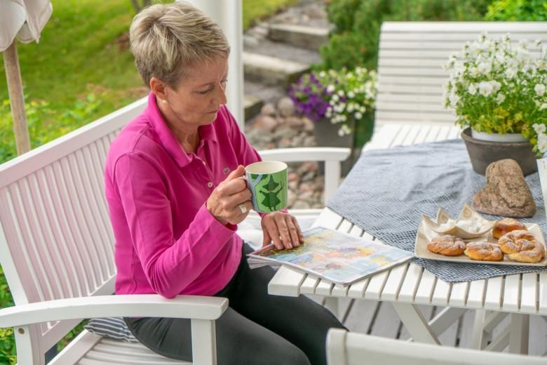 Eläkeikäinen nainen istuu terassilla kahvilla ja lukee lehteä.