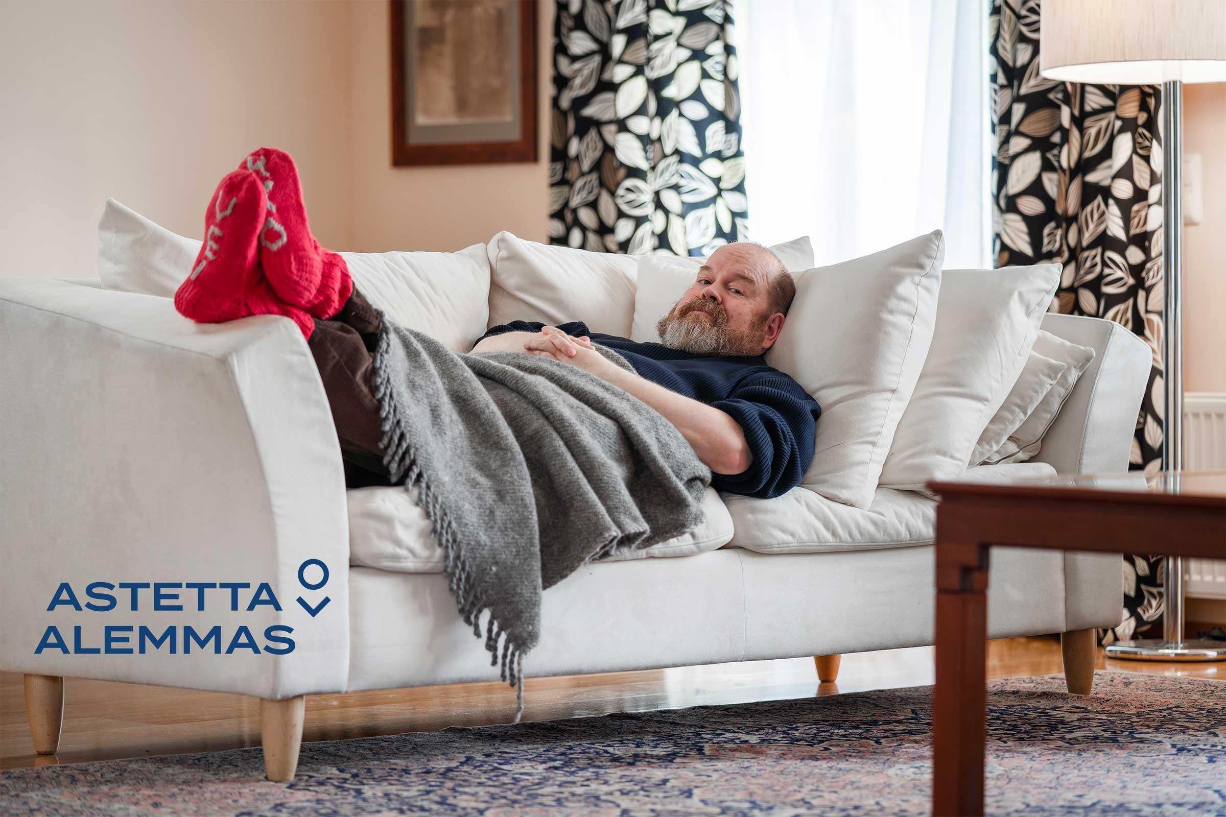 Astetta alemmas -kampanjan valokuva, jossa mies makaa sohvalla villasukat jalassa, Astetta alemmas -logo.