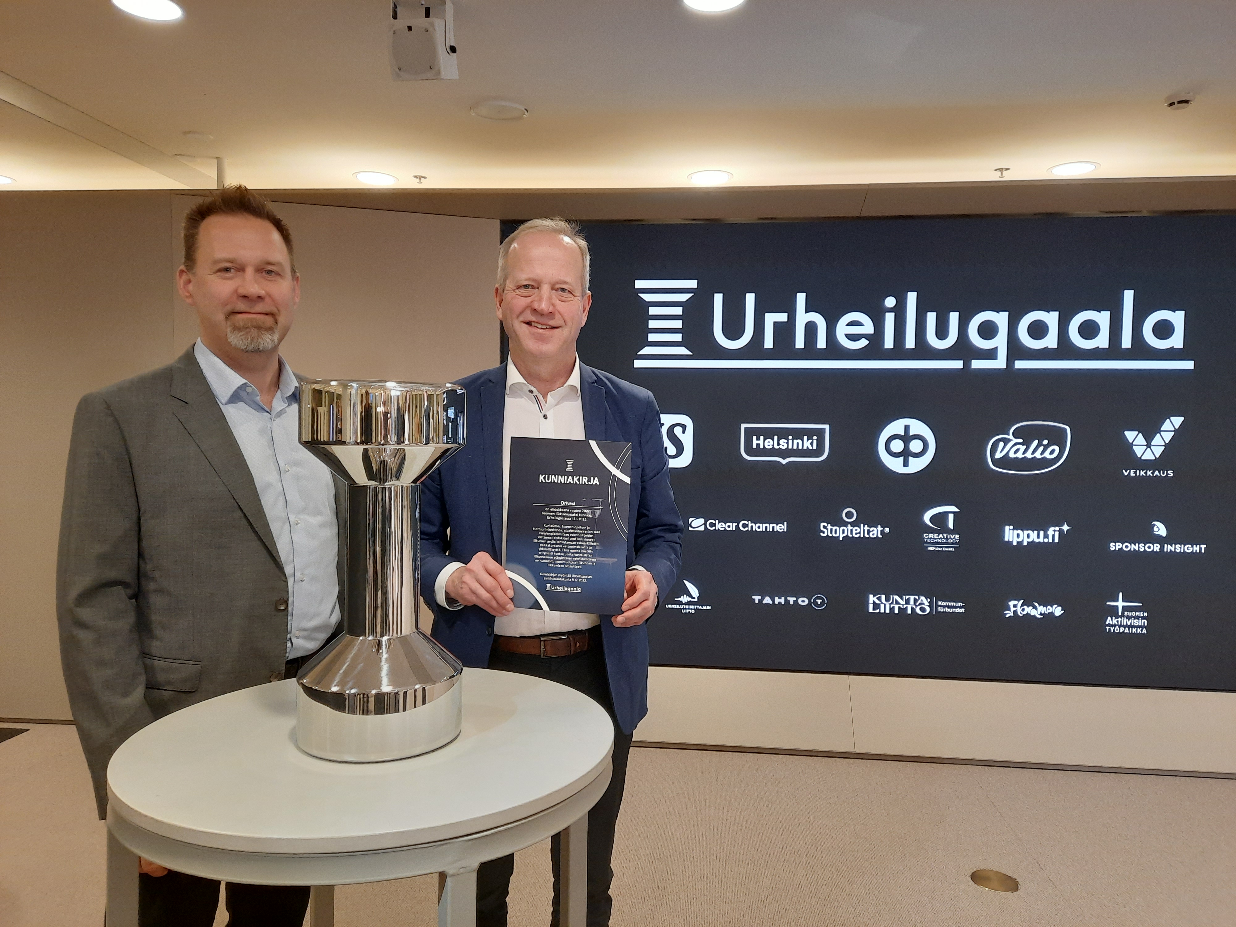 Liikuntapäällikkö Roope Marski ja kaupunginjohtaja Juha Kuusisto UNO-pokaalin ja kunniakirjan kanssa, taustalla Urheilugaalan kuvausseinä.